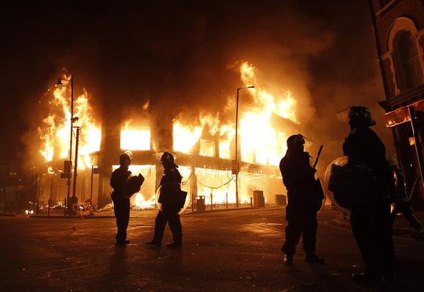 Police officers wearing riot gear stand in front of a burning building in Tottenham, north London August 7, 2011. Crowds attacked riot police and set two squad cars alight in north London on Saturday following a protest at the fatal shooting of a man by armed officers earlier in the week. 